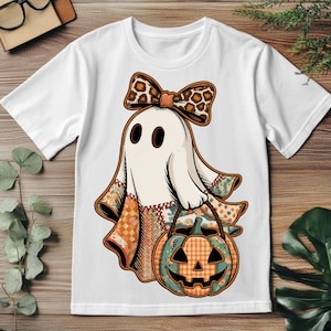 May include: A white short-sleeve t-shirt featuring a graphic of a ghost wearing a leopard print bow. The ghost is holding a jack-o'-lantern pumpkin bucket with a plaid pattern. The ghost's body is decorated with various autumn-themed patterns.