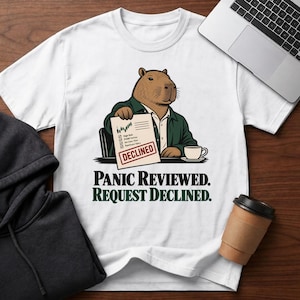 May include: A white t-shirt featuring a graphic of a brown capybara wearing a dark green shirt, sitting at a desk with a coffee cup. The capybara holds a document with a chart and a red stamp that reads DECLINED. Text below reads PANIC REVIEWED. REQUEST DECLINED.