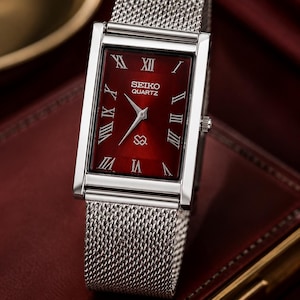 May include: A rectangular Seiko Quartz wristwatch featuring a deep red dial with silver-toned Roman numeral hour markers and silver hands. The watch has a polished silver-toned metal case and a flexible silver-toned stainless steel mesh strap.