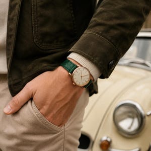 May include: A wristwatch with a round gold-tone case, a white dial with thin gold-tone hour markers, and a textured dark green leather strap. The watch is worn on a wrist, visible alongside a dark olive green jacket and beige trousers.