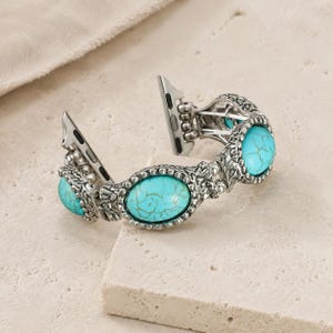 Peut inclure: Un bracelet de montre orné de pierres turquoise ovales avec des veines brunes, serties dans des maillons en métal argenté texturé et oxydé. Le bracelet comprend des connecteurs métalliques intégrés pour montre connectée, au fini détaillé.