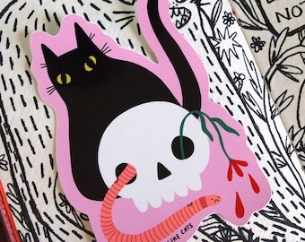 Könnte beinhalten: Ein rosa Aufkleber mit einer schwarzen Katze, die auf einem weißen Schädel sitzt. Die Katze hat gelbe Augen und der Schädel hat einen Wurm, der aus der Augenhöhle kommt. Der Aufkleber hat den Text "Ich mag Katzen" unten. Der Aufkleber befindet sich auf einer Seite eines Journals mit schwarzen Tintenzeichnungen von Blumen und Pflanzen.