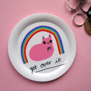 May include: White ceramic plate featuring a pink cat illustration with a rainbow behind it. The text "get over it" is written in black below the cat.