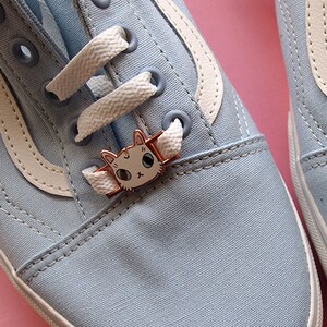 Op de afbeelding: Een paar lichtblauwe canvas schoenen met witte veters en een witte streep aan de zijkant. Elke schoen heeft een klein kattenbedeltje aan de veters.