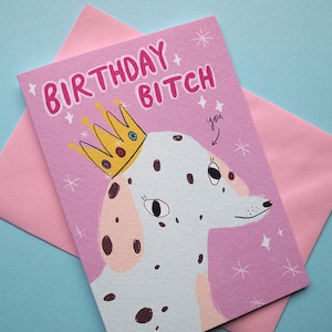 May include: A pink birthday card with the words "BIRTHDAY BITCH" in pink lettering. The card features a cartoon Dalmatian dog wearing a gold crown. A pink envelope is behind the card on a blue background.