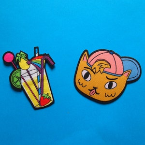 May include: Two embroidered iron-on patches. One is a yellow, orange, and pink cartoon of a glass of lemonade with a straw, a lime wedge, and a cherry. The other is a yellow cartoon cat wearing a pink and blue baseball cap.