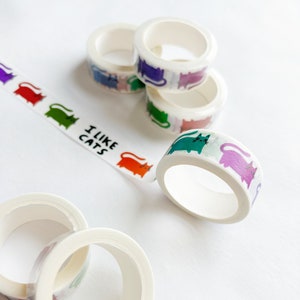 Peut inclure: Assortiment de rubans washi color&eacute;s avec un motif de chats r&eacute;p&eacute;t&eacute;. Les rubans sont enroul&eacute;s et empil&eacute;s sur une surface blanche. Un ruban porte l'inscription "J'aime les chats".