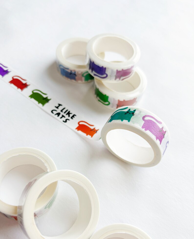 Peut inclure: Assortiment de rubans washi color&eacute;s avec un motif de chats r&eacute;p&eacute;t&eacute;. Les rubans sont enroul&eacute;s et empil&eacute;s sur une surface blanche. Un ruban porte l'inscription "J'aime les chats".