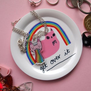 May include: A white ceramic dish with a pink cat and rainbow design. The dish has the text "get over it" written on it. The dish is surrounded by various jewellery pieces, including a silver necklace with a pink cat charm.