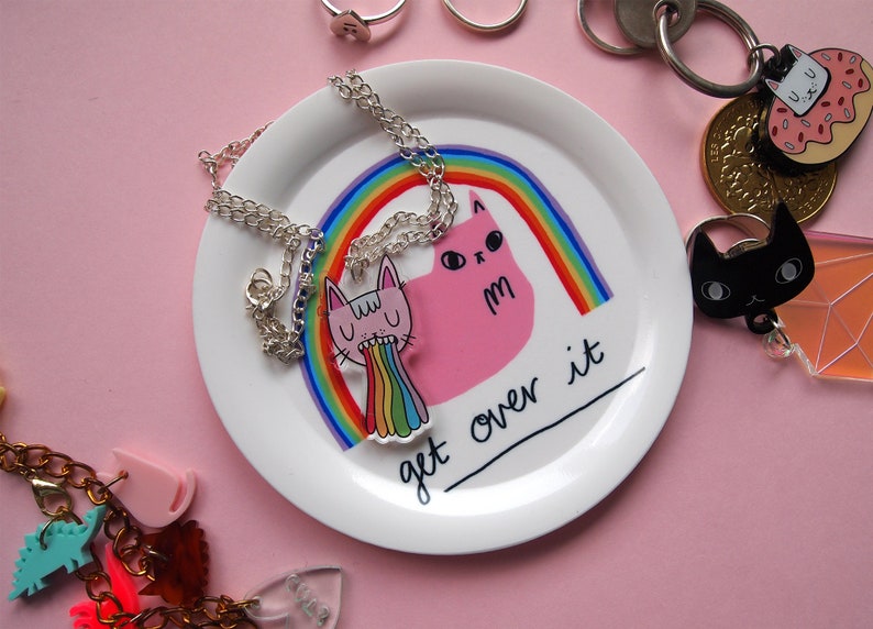 May include: A white ceramic dish with a pink cat and rainbow design. The dish has the text "get over it" written on it. The dish is surrounded by various jewellery pieces, including a silver necklace with a pink cat charm.