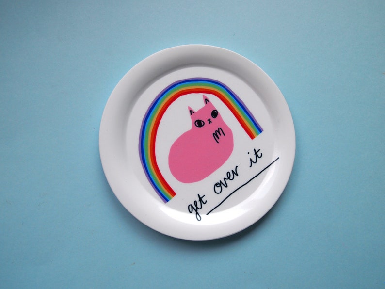 May include: White ceramic plate featuring a pink cat sitting on a rainbow with the text "get over it".