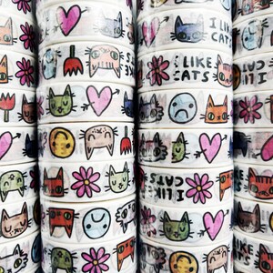 Peut inclure: Une collection de rubans washi blancs avec des illustrations color&eacute;es de chats, de c&oelig;urs, de fleurs et de visages souriants. Certains rubans ont du texte qui dit "J'aime les chats" et "J'aime les chats".