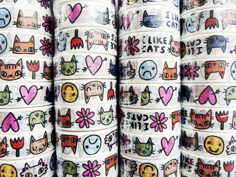 Peut inclure: Une collection de rubans washi blancs avec des illustrations color&eacute;es de chats, de c&oelig;urs, de fleurs et de visages souriants. Certains rubans ont du texte qui dit "J'aime les chats" et "J'aime les chats".