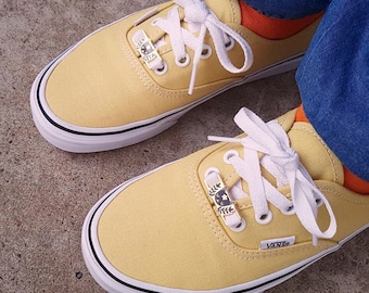 Op de afbeelding: Een paar gele Vans sneakers van canvas met witte veters en zwarte trim. De schoenen hebben decoratieve bedels aan de veters.