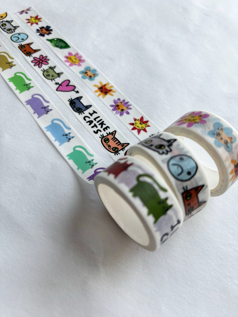 Peut inclure: Quatre rouleaux de ruban adh&eacute;sif washi color&eacute; avec des motifs de chats. Le ruban pr&eacute;sente des chats avec diff&eacute;rentes expressions, notamment un c&oelig;ur, un poisson et les mots "J'aime les chats".