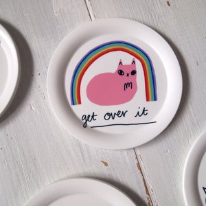 May include: A white ceramic plate featuring a pink cat sitting beneath a rainbow. The cat has a grumpy expression and the text "get over it" is written below the rainbow.
