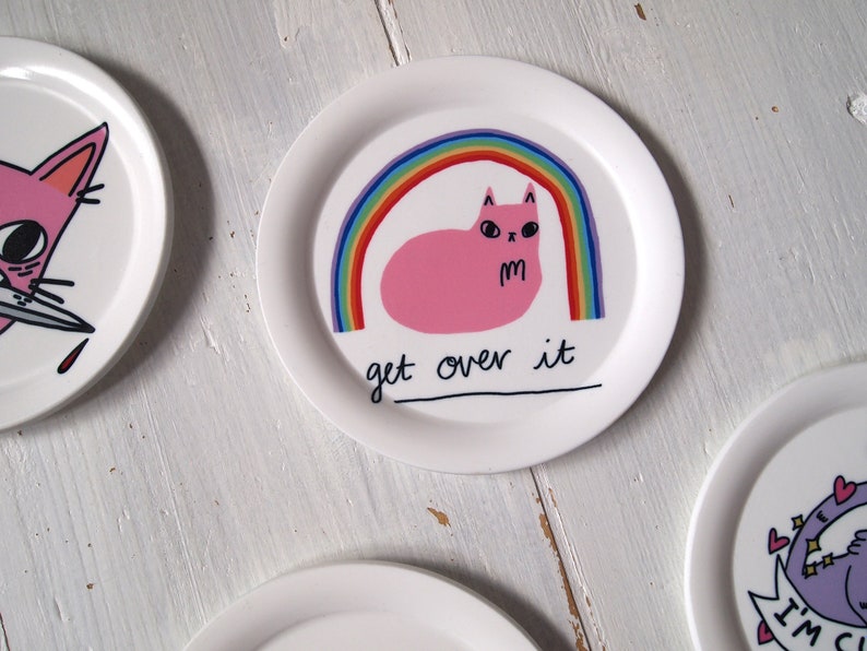 May include: A white ceramic plate featuring a pink cat sitting beneath a rainbow. The cat has a grumpy expression and the text "get over it" is written below the rainbow.