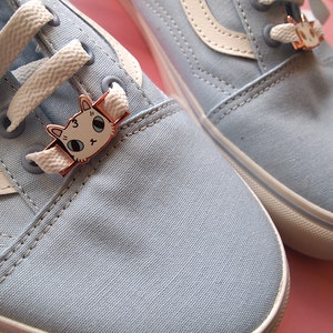 Op de afbeelding: Een paar lichtblauwe canvas schoenen met witte veters en een witte streep. Elke schoen heeft een klein kattenbedeltje aan de veters.