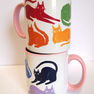 May include: Two white ceramic mugs with pink handles. The mugs are stacked on top of each other. The top mug has a design of rainbow colored cats. The bottom mug has a design of purple, blue, and black cats.