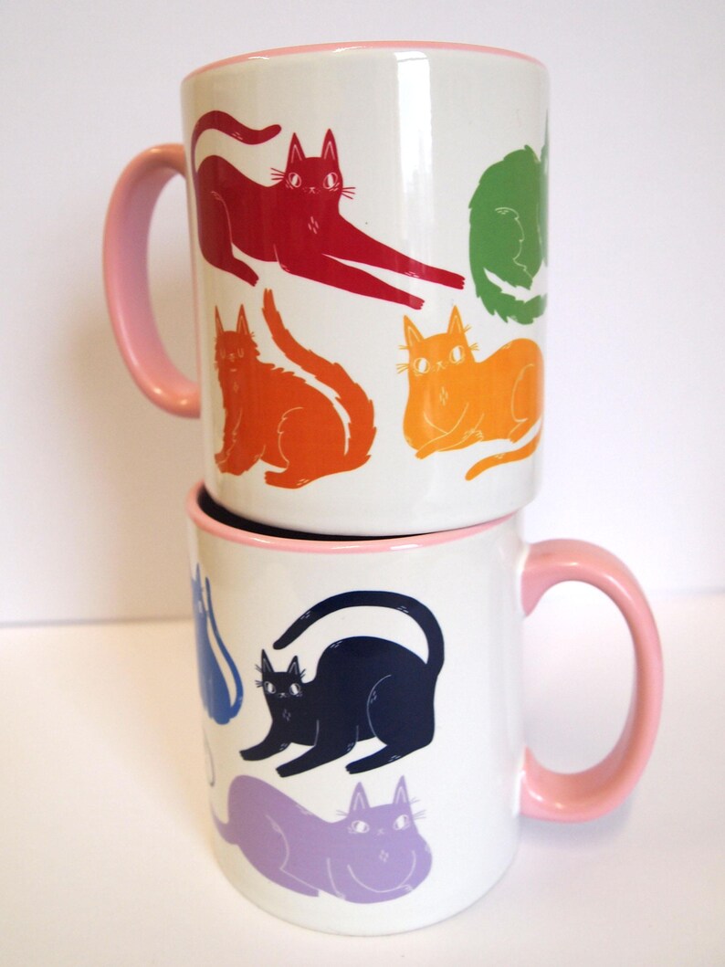 May include: Two white ceramic mugs with pink handles. The mugs are stacked on top of each other. The top mug has a design of rainbow colored cats. The bottom mug has a design of purple, blue, and black cats.