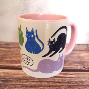 May include: A white ceramic mug with a pink rim and handle. The mug features a colorful pattern of cats with the text "I like cats" on the front.