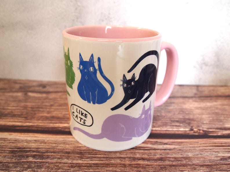 May include: A white ceramic mug with a pink rim and handle. The mug features a colorful pattern of cats with the text "I like cats" on the front.