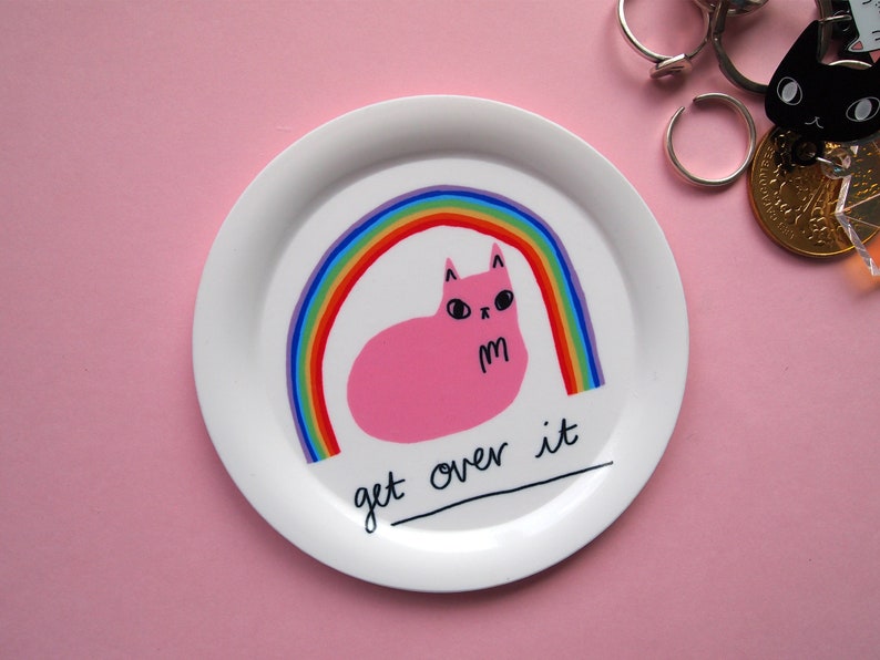 May include: White ceramic plate featuring a pink cat illustration with a rainbow behind it. The text "get over it" is written in black below the cat.