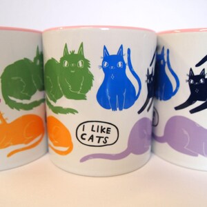 May include: Three white ceramic mugs with pink rims, each featuring a different colorful cat illustration. The middle mug has the text "I like cats" printed on it.