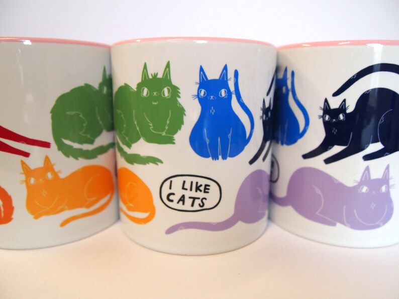 May include: Three white ceramic mugs with pink rims, each featuring a different colorful cat illustration. The middle mug has the text "I like cats" printed on it.