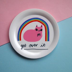 May include: White ceramic plate featuring a pink cat lying under a rainbow with the text "get over it".