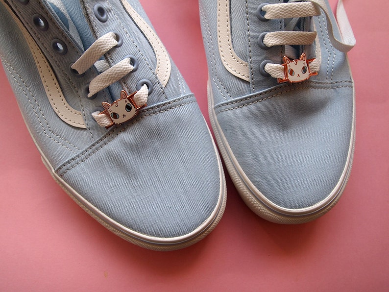 Op de afbeelding: Een paar lichtblauwe canvas schoenen met witte veters en een witte streep aan de zijkant. Elke schoen heeft een klein kattenbedeltje aan de veters.