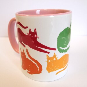 May include: A white ceramic mug with a pink rim features a colorful design of five cats in various poses. The cats are red, orange, yellow, green, and blue.