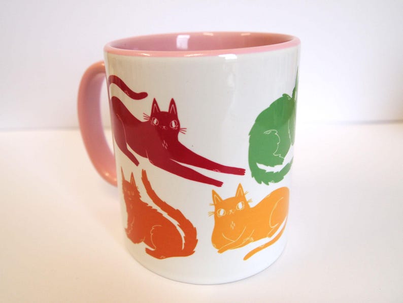 May include: A white ceramic mug with a pink rim features a colorful design of five cats in various poses. The cats are red, orange, yellow, green, and blue.