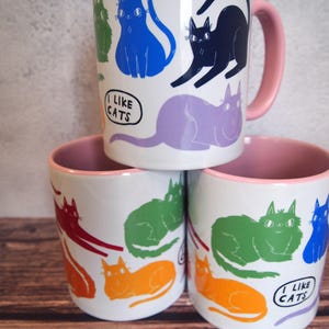 May include: Three white ceramic mugs with pink rims. Each mug features a different colorful cat design and the text "I like cats".
