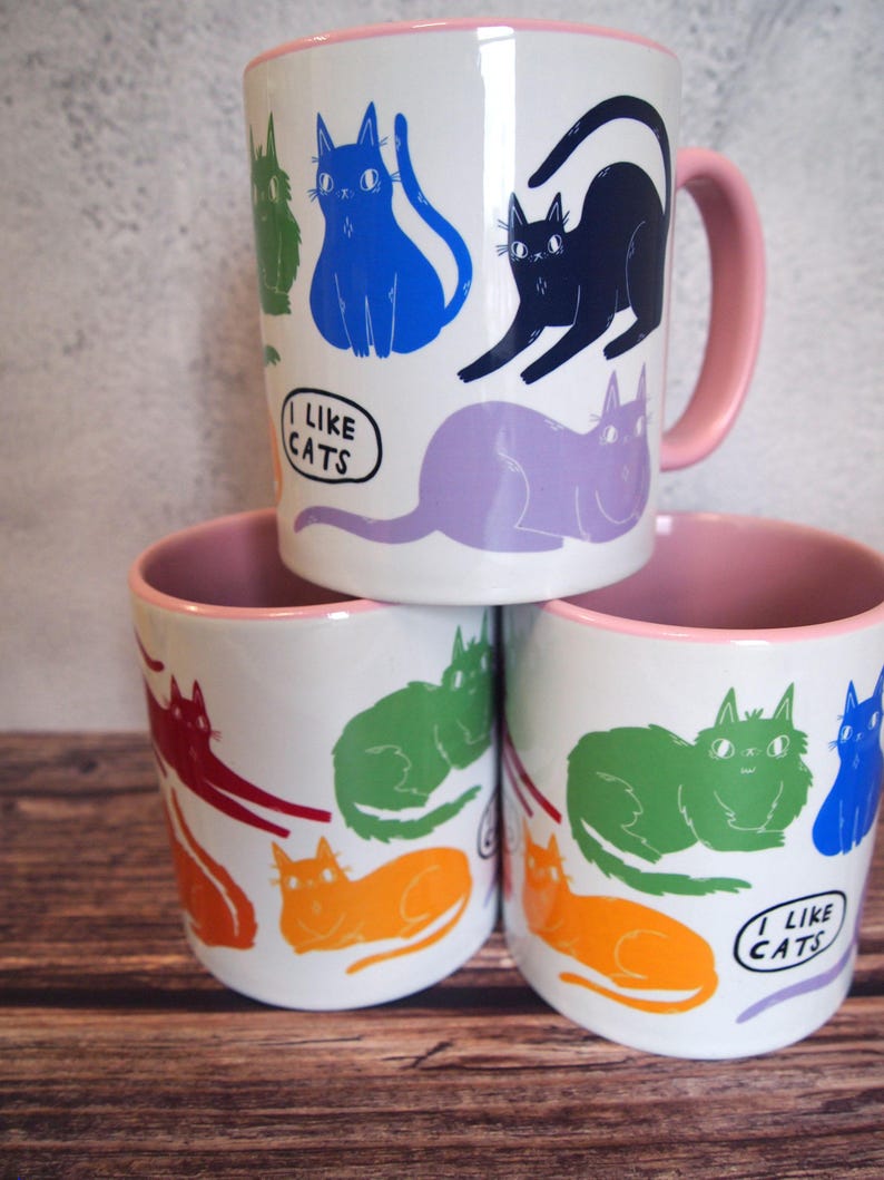 May include: Three white ceramic mugs with pink rims. Each mug features a different colorful cat design and the text "I like cats".