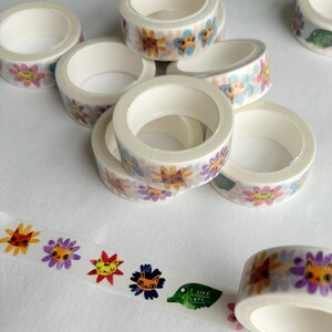Peut inclure: Un ensemble de six rouleaux de ruban adh&eacute;sif washi avec des motifs floraux color&eacute;s et des visages de chats. Le ruban est blanc avec un fond transparent et les motifs sont imprim&eacute;s sur le ruban. Le ruban est enroul&eacute; et empil&eacute; les uns sur les autres. Un rouleau de ruban est d&eacute;roul&eacute; et montre le motif plus clairement. Le ruban est &eacute;tiquet&eacute; "J'aime les chats".