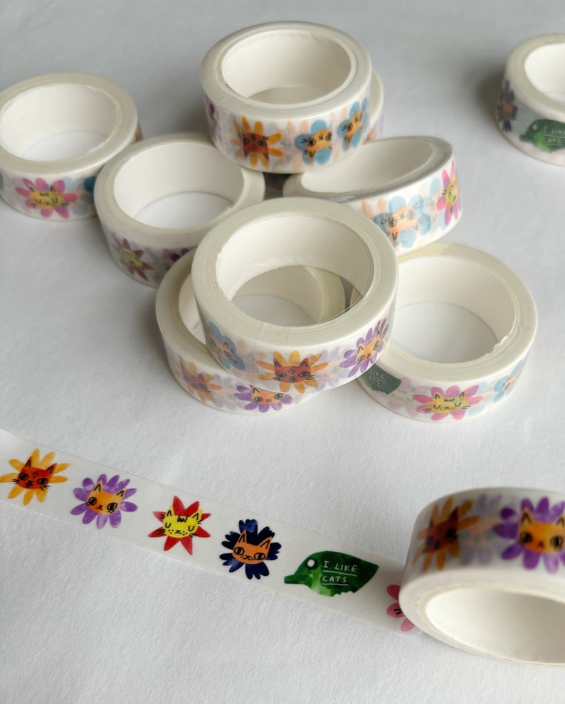Peut inclure: Un ensemble de six rouleaux de ruban adh&eacute;sif washi avec des motifs floraux color&eacute;s et des visages de chats. Le ruban est blanc avec un fond transparent et les motifs sont imprim&eacute;s sur le ruban. Le ruban est enroul&eacute; et empil&eacute; les uns sur les autres. Un rouleau de ruban est d&eacute;roul&eacute; et montre le motif plus clairement. Le ruban est &eacute;tiquet&eacute; "J'aime les chats".