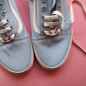 Op de afbeelding: Een paar lichtblauwe canvas sneakers met witte veters en schoenbedels in de vorm van een kat. De schoenen staan op een roze achtergrond.