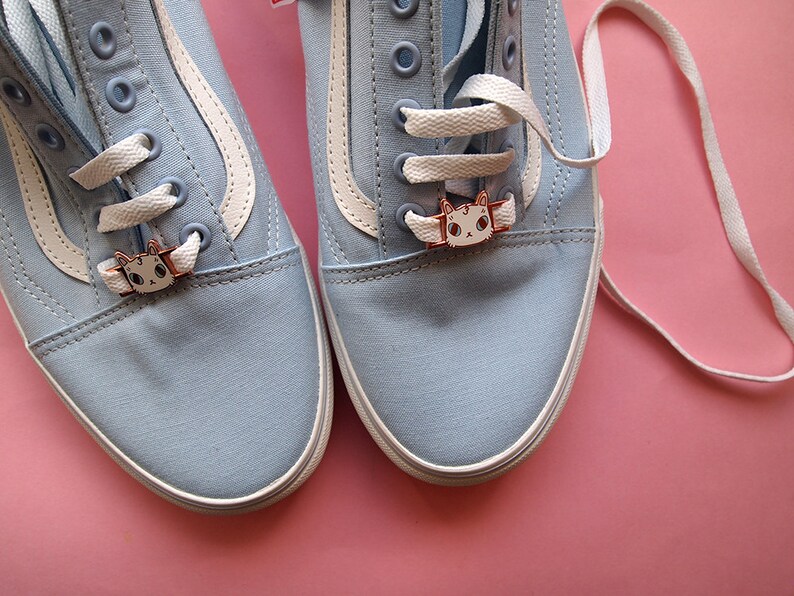 Op de afbeelding: Een paar lichtblauwe canvas sneakers met witte veters en schoenbedels in de vorm van een kat. De schoenen staan op een roze achtergrond.