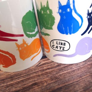 May include: Two white ceramic mugs with colorful cat illustrations. The mug on the left has red, orange, green, and yellow cats. The mug on the right has blue, purple, and black cats. The mug on the right has the text "I like cats" in a speech bubble.