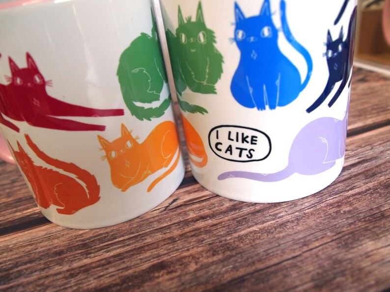 May include: Two white ceramic mugs with colorful cat illustrations. The mug on the left has red, orange, green, and yellow cats. The mug on the right has blue, purple, and black cats. The mug on the right has the text "I like cats" in a speech bubble.