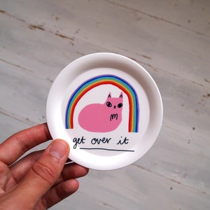 May include: A white ceramic coaster featuring a pink cat sitting under a rainbow with the text "get over it".