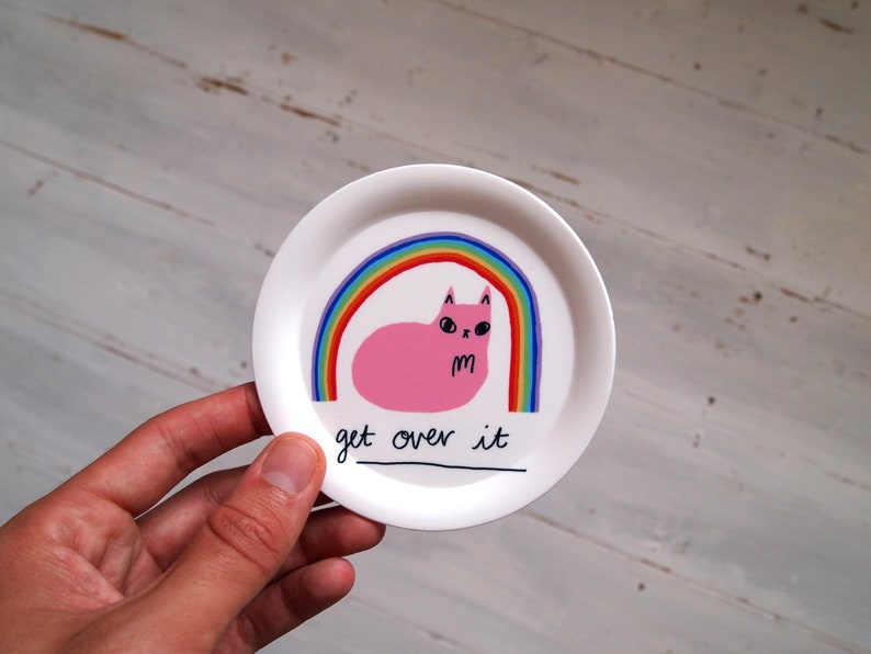 May include: A white ceramic coaster featuring a pink cat sitting under a rainbow with the text "get over it".