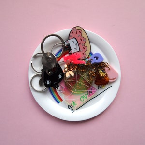 May include: A white ceramic dish with a rainbow design and the text "get over it" features a variety of colourful keychains, including a black cat, a pink donut, a pink heart, and a gold chain.