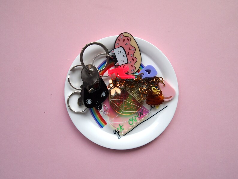May include: A white ceramic dish with a rainbow design and the text "get over it" features a variety of colourful keychains, including a black cat, a pink donut, a pink heart, and a gold chain.
