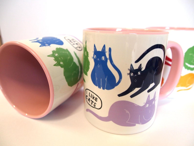 May include: Four pink and white ceramic mugs with a cat design. Each mug features a different colored cat with the text "I like cats" on one mug.