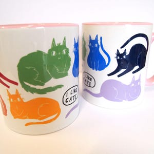 May include: Two white ceramic mugs with pink rims. Both mugs feature a colorful cat design and the text "I like cats".