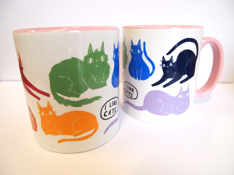 May include: Two white ceramic mugs with pink rims. Both mugs feature a colorful cat design and the text "I like cats".