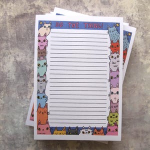 Puede incluir: Una pila de blocs de notas rayados con un borde de gatos coloridos. El bloc de notas superior tiene el texto "DO THE THING!" en azul. El borde presenta varios gatos de dibujos animados en tonos rosa, naranja, azul y verde.