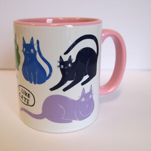 May include: A white ceramic mug with a pink rim and handle. The mug features a colorful illustration of cats with the text "I like cats" in a speech bubble.